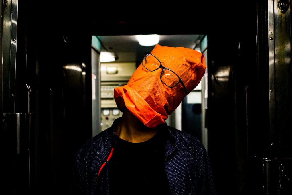 figure in a hallway with a orange bag on head and glasses on top of the bag, head titled to the side, showing the absurdity of art cliches